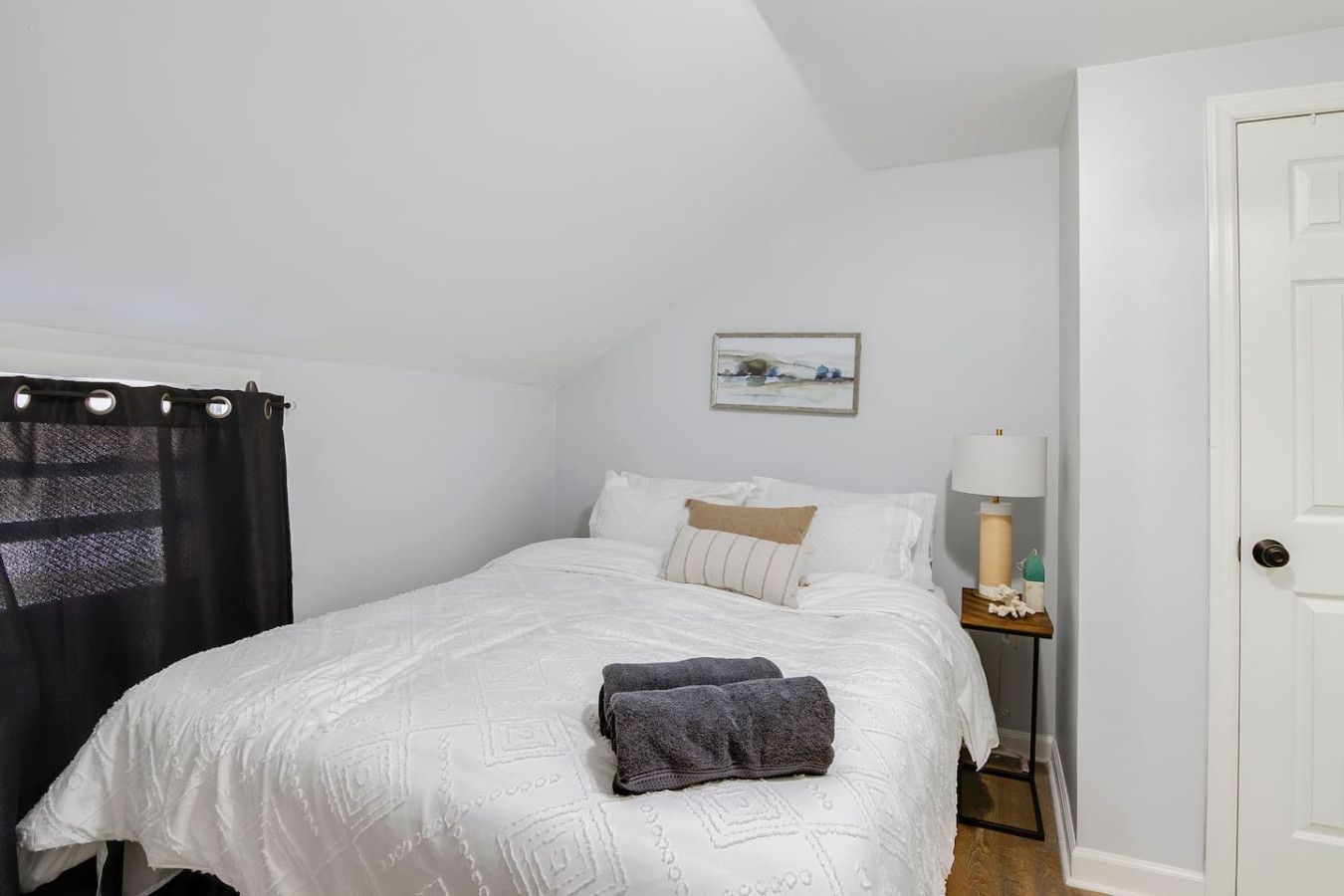 Quarto com cama, mesa de cabeceira, luminária, porta e janela com cortina.