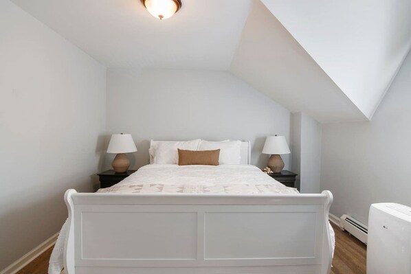 A bedroom with a white bed, two bedside tables with lamps, a sloped ceiling, and a radiator.