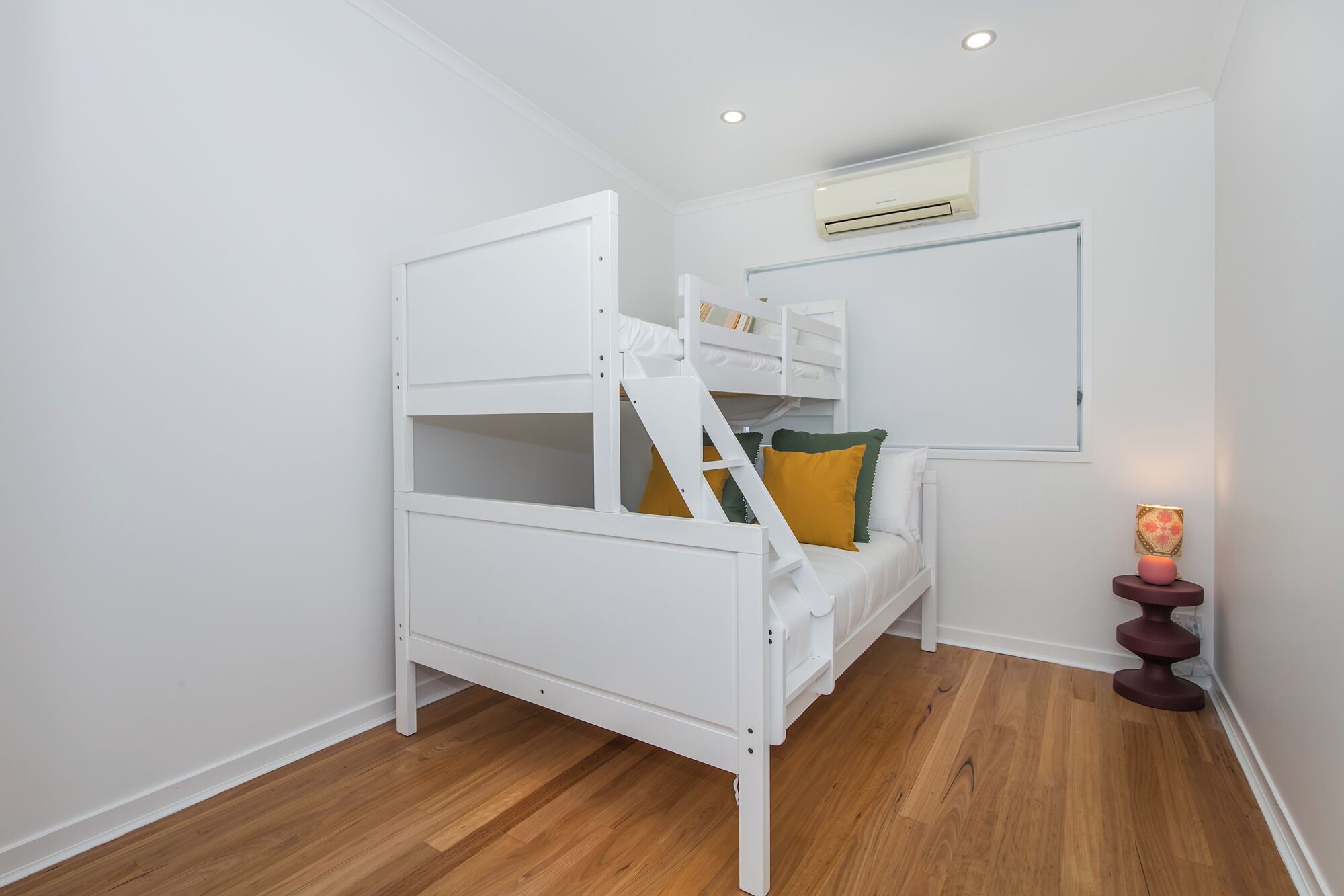 Una habitación pequeña y blanca con una cama litera, suelo de madera, un equipo de aire acondicionado y una pequeña mesa con una lámpara.