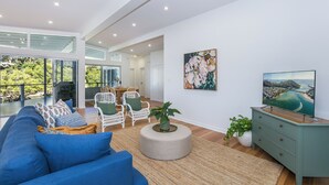 Uma sala de estar espaçosa com um sofá azul, cadeiras de vime branco, um banquinho redondo e uma cômoda azul-esverdeada. Um quadro grande e uma televisão estão na parede.