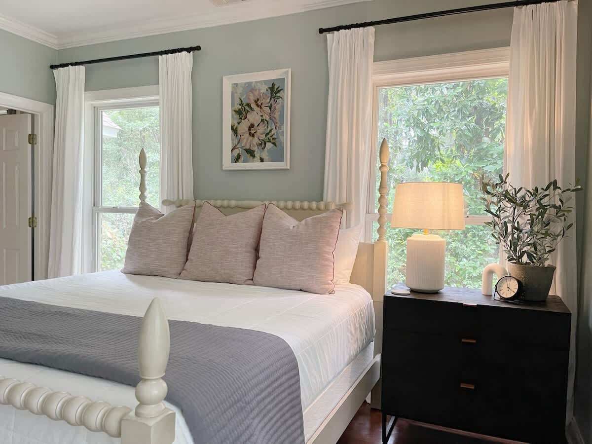 Un dormitorio con cama, mesita de noche, lámpara, reloj y ventana con cortinas.