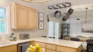 A kitchen with wooden cabinets, a central island with a bowl of fruit, a hanging pot rack, and a dining area with chairs and a table. - Savannah Home with Modern Airstream, Near Tybee (Savannah)