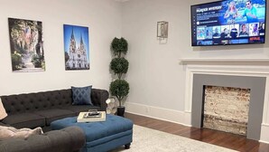 Ein Wohnzimmer mit einem dunkelgrauen Sofa, einem blauen Sessel, einem kleinen runden Tisch und einem Kamin. An der Wand hängen zwei gerahmte Bilder und über dem Kamin ist ein Fernseher angebracht.