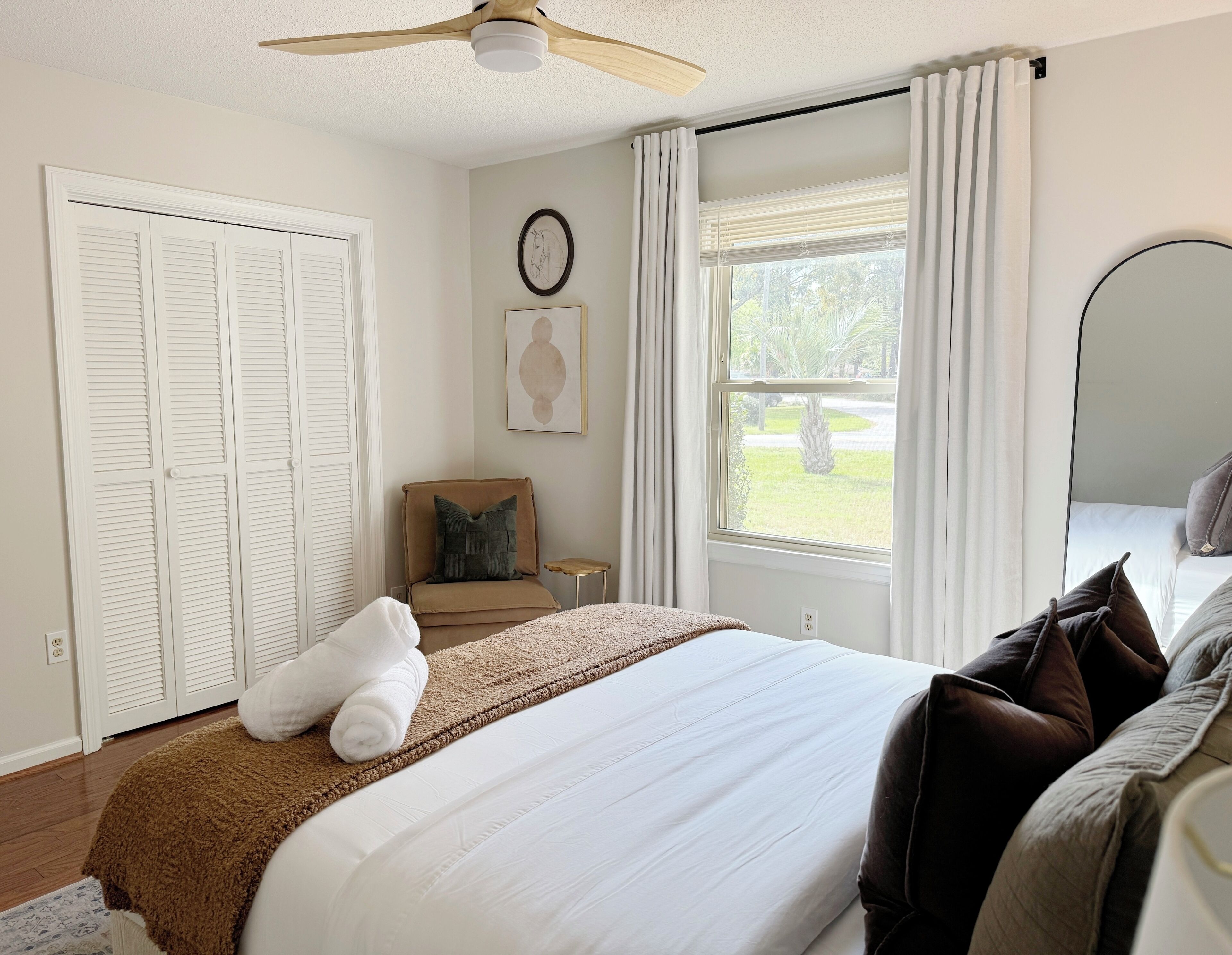 Une chambre à coucher comprenant un lit, une chaise, une fenêtre avec des rideaux, un ventilateur de plafond et un miroir fixé au mur.