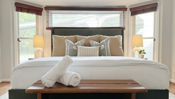 A neatly made bed with a rolled towel on a wooden bedside table, a headboard with multiple pillows, and two bedside lamps. - Poolside Family Haven Near Beach w/ Game Room (Savannah)