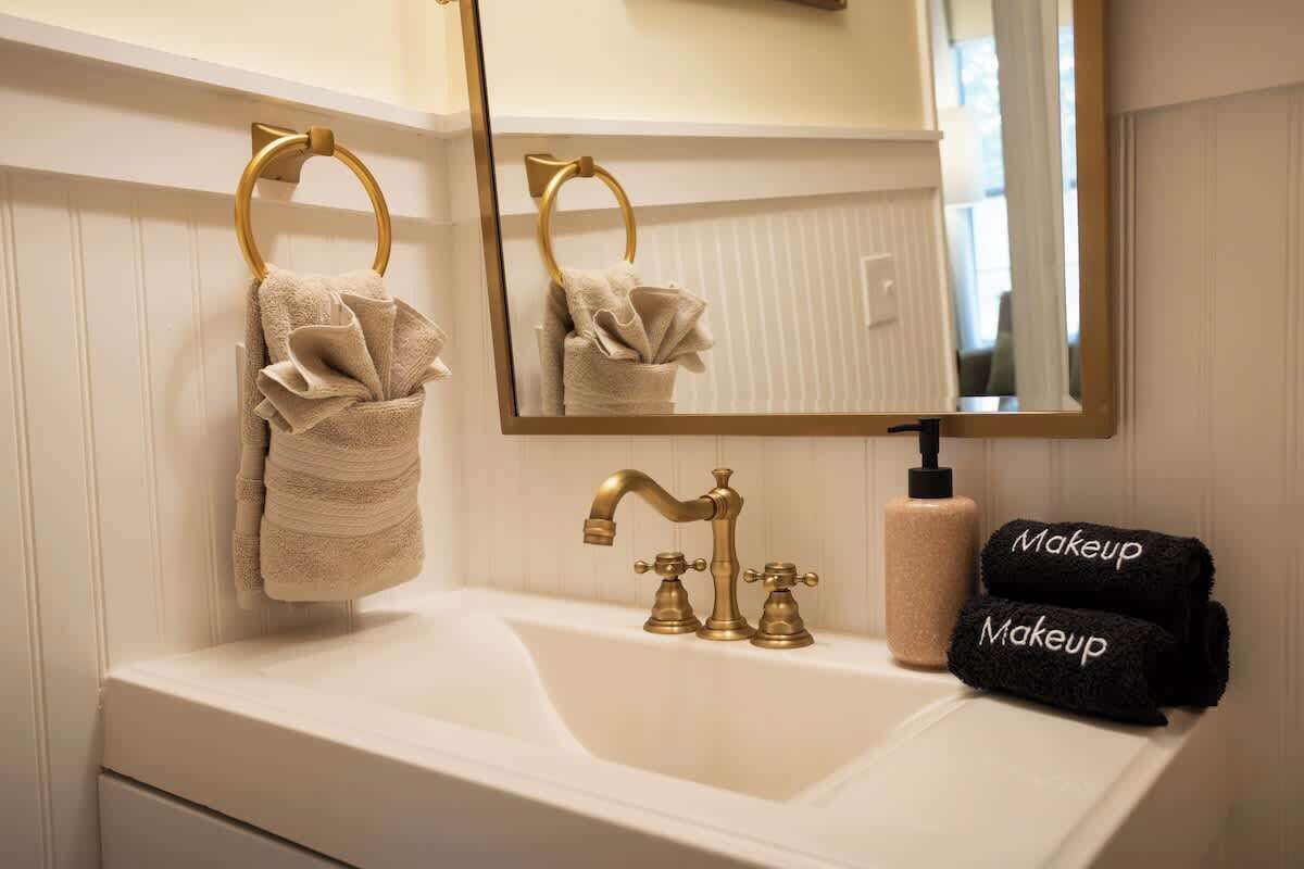 A bathroom with a white sink, gold faucets, a gold towel ring, a mirror, and a bottle of shampoo.