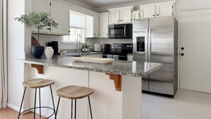 Una cocina moderna con armarios blancos, electrodomésticos de acero inoxidable, encimera de granito y dos taburetes de madera.