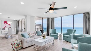 A modern living room with a white sofa, teal armchairs, and a coffee table. A ceiling fan is visible, and the room has large windows offering a view of the ocean.