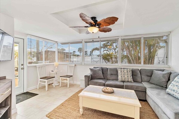 A spacious living room with a grey sofa, a coffee table, a ceiling fan, and large windows with blinds.