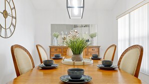 Ein Esszimmer mit einem Holztisch, sechs Stühlen, einer zentralen Vase mit Grünpflanzen und einem großen Spiegel über dem Tisch.