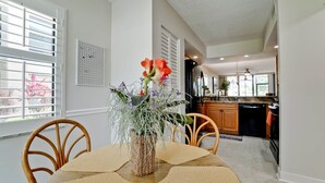 A dining area with a table set for two, a vase with flowers, and a view into the kitchen.