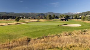 Un parcours de golf avec un ciel dégagé, des montagnes lointaines et quelques bâtiments en arrière-plan.