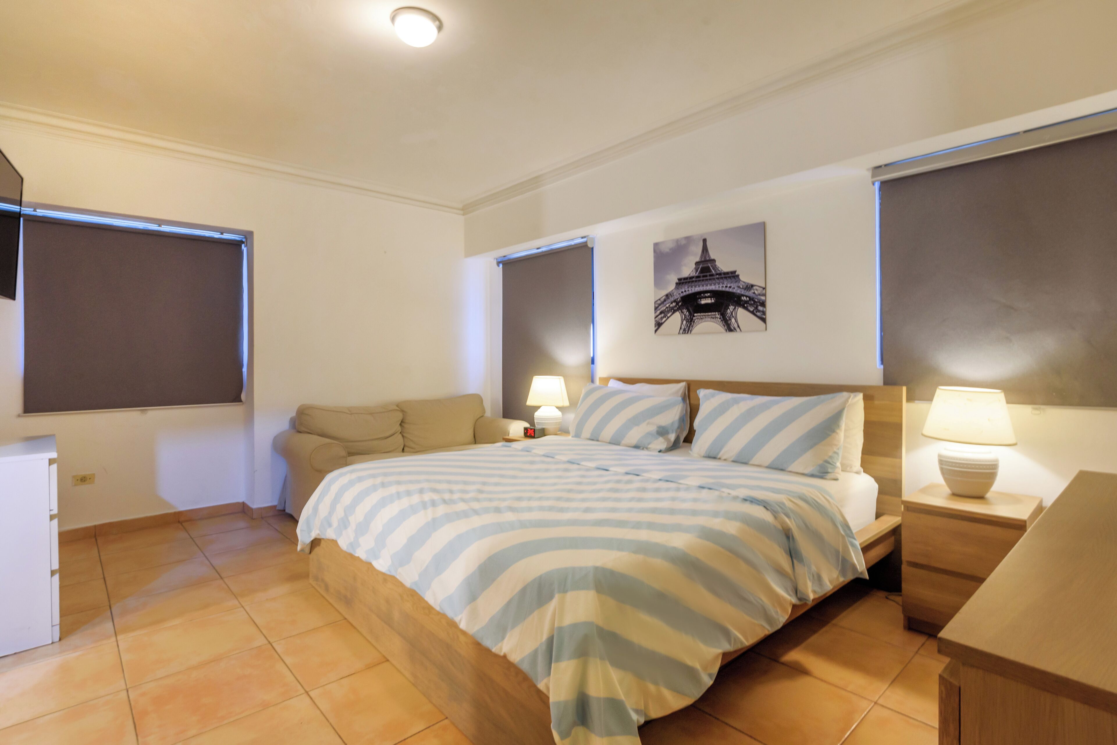 Un dormitorio con cama, mesitas de noche, lámpara, un cuadro en la pared y una ventana.