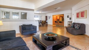 A spacious living room with a dark sofa, a coffee table, and a gray couch. There is a kitchen area in the background with wooden cabinets and a dining table.