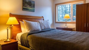 A neatly made bed with a wooden headboard, two bedside lamps, a painting on the wall, and a window with a view of trees. - Ski In/Out Right to the Elk Chairlift (Fernie)