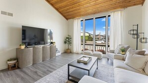 Een moderne woonkamer met een grote televisie, houten plafond en uitzicht op de stad door grote ramen.