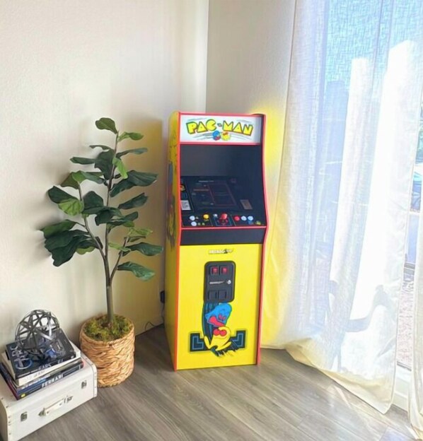 Una macchina arcade Pac-Man in un angolo della stanza, con una pianta in vaso e una pila di libri.