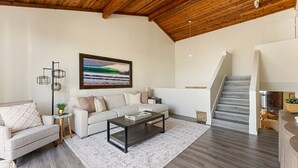 Een ruime woonkamer met een hoog houten plafond, een witte bank, een donkere salontafel en een trap die naar een hoger niveau leidt.