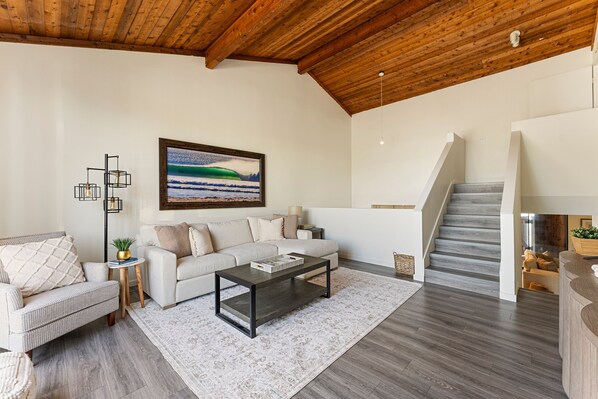 A spacious living room with a high wooden ceiling, a white sofa, a dark coffee table, and a staircase leading to another level. - The Blue Pearl - 2 minute walk to the beach! (Del Mar)