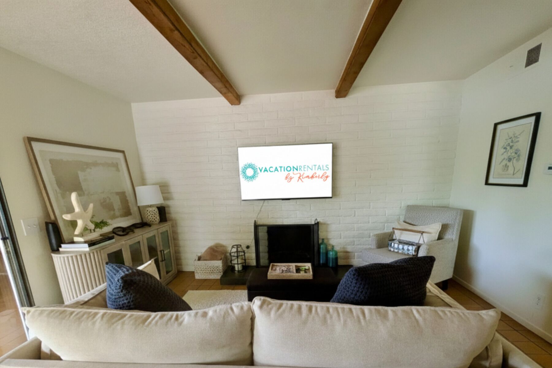 Una sala de estar con pared de ladrillo blanco, un televisor de pantalla plana sobre la chimenea, un sofá color beige con cojines oscuros y una viga de techo de madera.