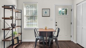 A dining area with a wooden table, two chairs, a metal shelf with decorative items, a window with blinds, and a door with a view of greenery. - Charming villa steps from the water in beautiful Pensacola (Pensacola)