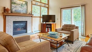 A spacious living room with a fireplace, a television, and a large window offering a view of greenery. - Overlook Poolside Getaway Heated Pool (May - Oct 20th) (East Falmouth)