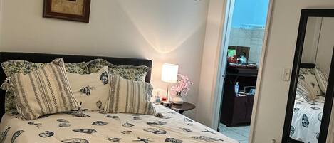 A bedroom with a bed, a ceiling fan, a nightstand with a lamp, a framed picture on the wall, and a door leading to another room.