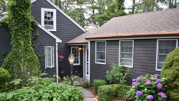 Une maison à l’extérieur d’un gris foncé, avec une porte blanche et un petit jardin devant, un chemin menant à la porte.