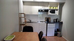 A compact kitchen with white cabinets, a microwave, a sink, and a small refrigerator. There is a wooden dining table with chairs and a green bowl on it.