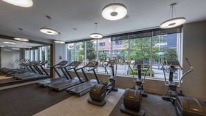 A gym with treadmills, elliptical machines, and free weights, large windows with a view of buildings and trees. - Tysons 2BR | Balcony, Pool & Fitness (McLean)