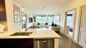 A modern kitchen with a central island, stainless steel appliances, pendant lighting, and a view of the living area with a sofa and a coffee table. - Tysons 2BR | Balcony, Pool & Fitness (McLean)