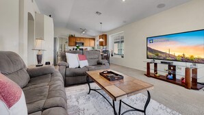 A spacious living room with a gray sofa, a coffee table, and a TV on a stand. - 3BR w private yard, grill, garden view (San Tan Valley)