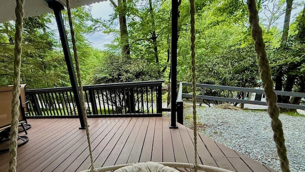 Una terraza de madera con una mecedora colgante, rodeada de árboles y un sendero de grava.