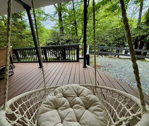 A wooden deck with a hanging chair, surrounded by trees and a gravel path.