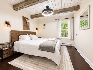 A bedroom with a wooden headboard, a bed with white bedding, a grey throw, a bedside table with a lamp, a window with curtains, and a view of trees.