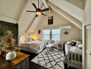 A cozy attic bedroom with a sloped ceiling, a ceiling fan, a large window, a bed, a chair, a crib, and a decorative plant.