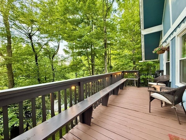 A wooden deck with a railing, wicker furniture, and a view of dense green trees.