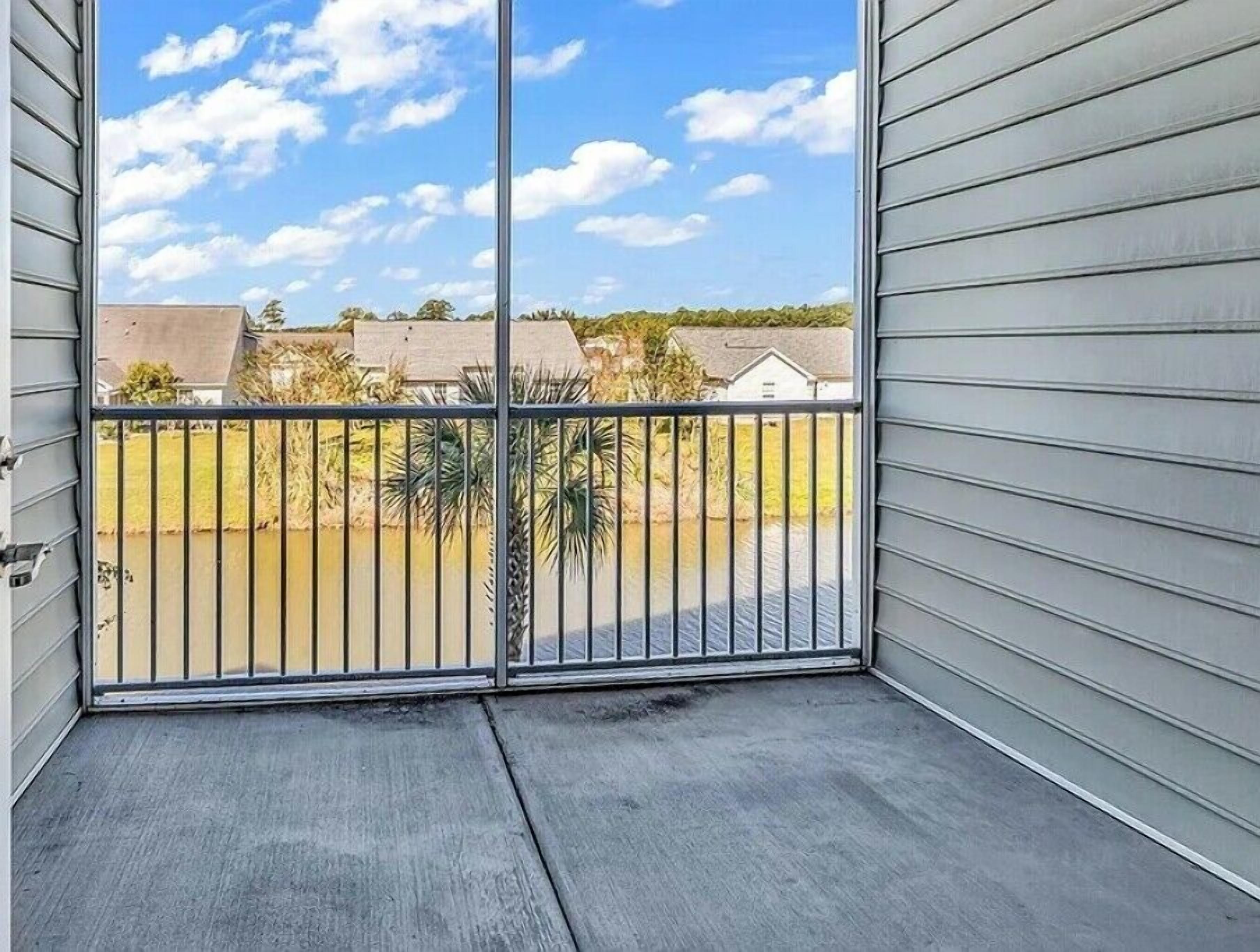 A balcony with a metal railing, a concrete floor, and a view of houses and trees in the background.