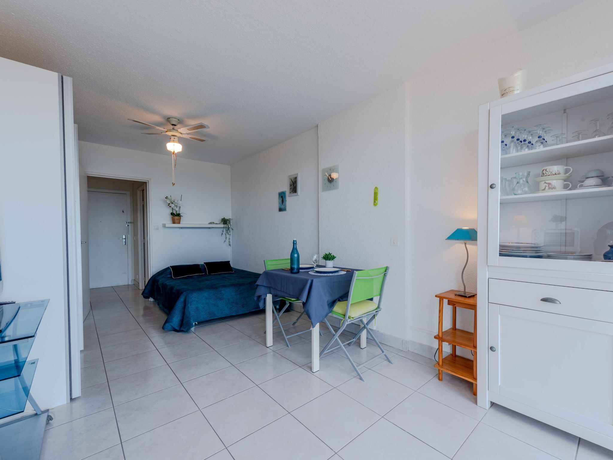Quarto compacto com cama, mesa de jantar com cadeiras, ventilador de teto e armário com louças.