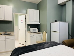 A compact hotel room with a kitchenette, a bed, a chair, and a refrigerator.