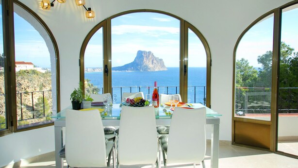 A dining area with a table set for two, large arched windows offering a view of the sea and a mountain, and a potted plant on the table. - Gasparet by Interhome (Calpe/Calp)