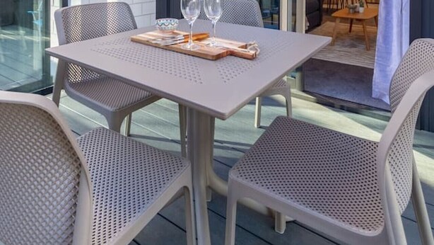Un área de comedor al aire libre con una mesa, sillas, barandilla de cristal y vista hacia el interior del edificio.
