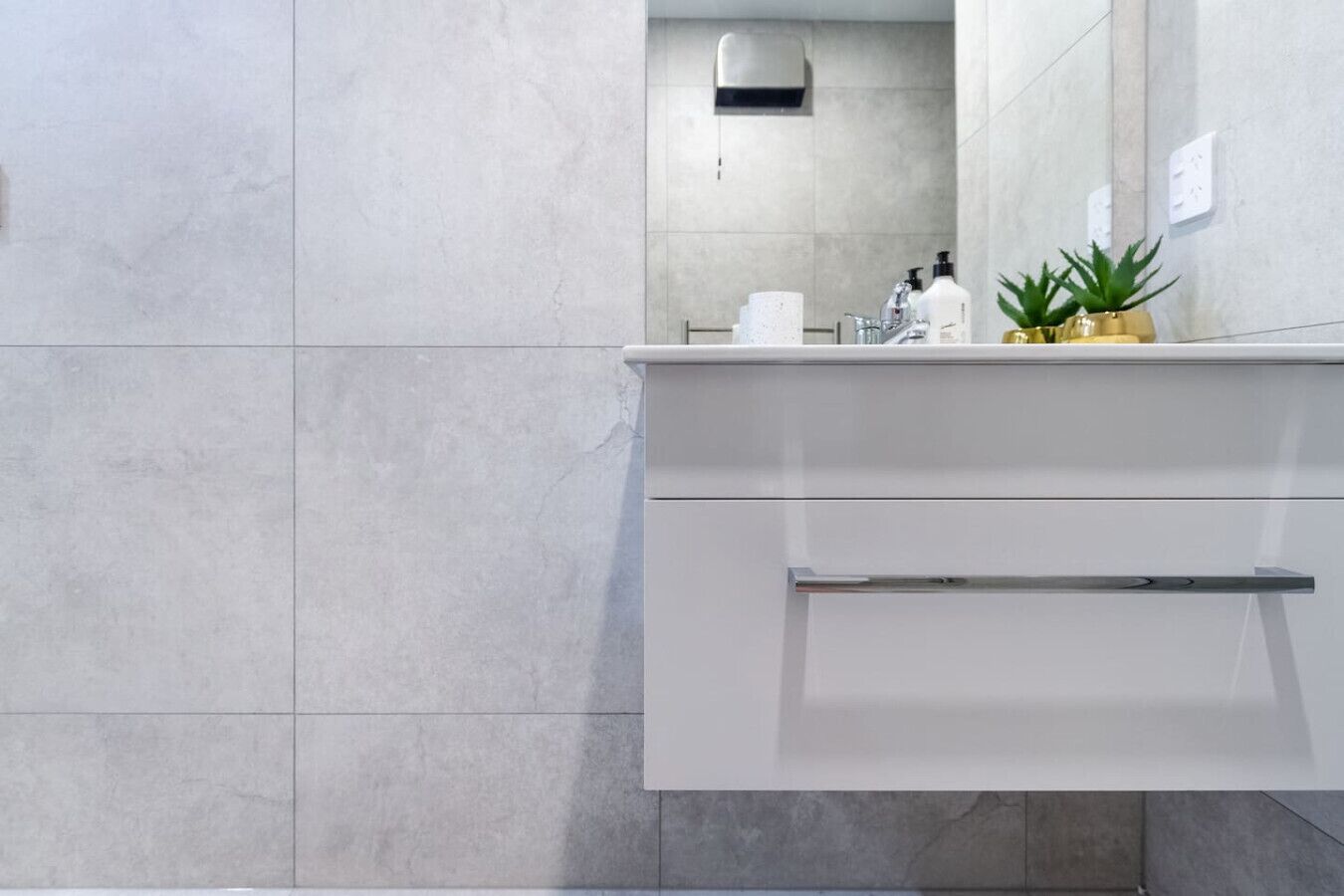 Un baño moderno con baldosas grises, un tocador blanco, un espejo y una planta pequeña en la encimera.