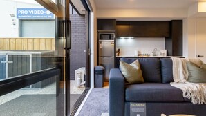 Una sala de estar moderna con sofá cama, una zona de cocina al fondo y una puerta de vidrio que da al exterior.