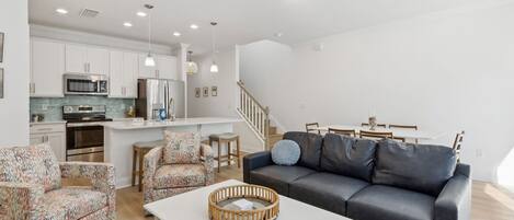A modern living room with a black leather sofa, patterned armchairs, and a white coffee table. A staircase leads to a dining area with wooden chairs.