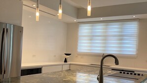 Una cocina moderna con una isla central, electrodomésticos de acero inoxidable, iluminación colgante y una ventana con persianas.