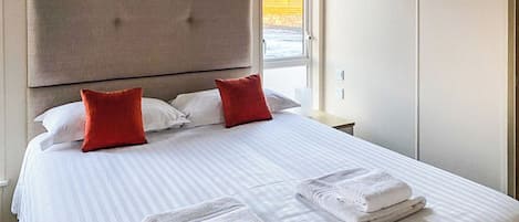 Une chambre d’hôtel avec un grand lit, deux oreillers rouges, une tête de lit, une table de chevet et une fenêtre donnant sur un beau paysage.