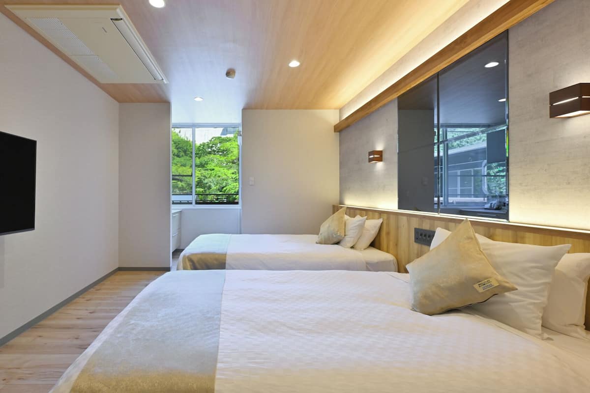 A hotel room with two beds, a flat-screen TV, and a large window offering a view of greenery.