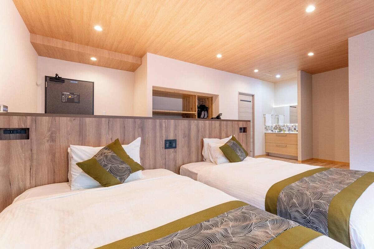 A hotel room with two beds, a wooden headboard, a built-in wardrobe, a mirror, and a small kitchenette.
