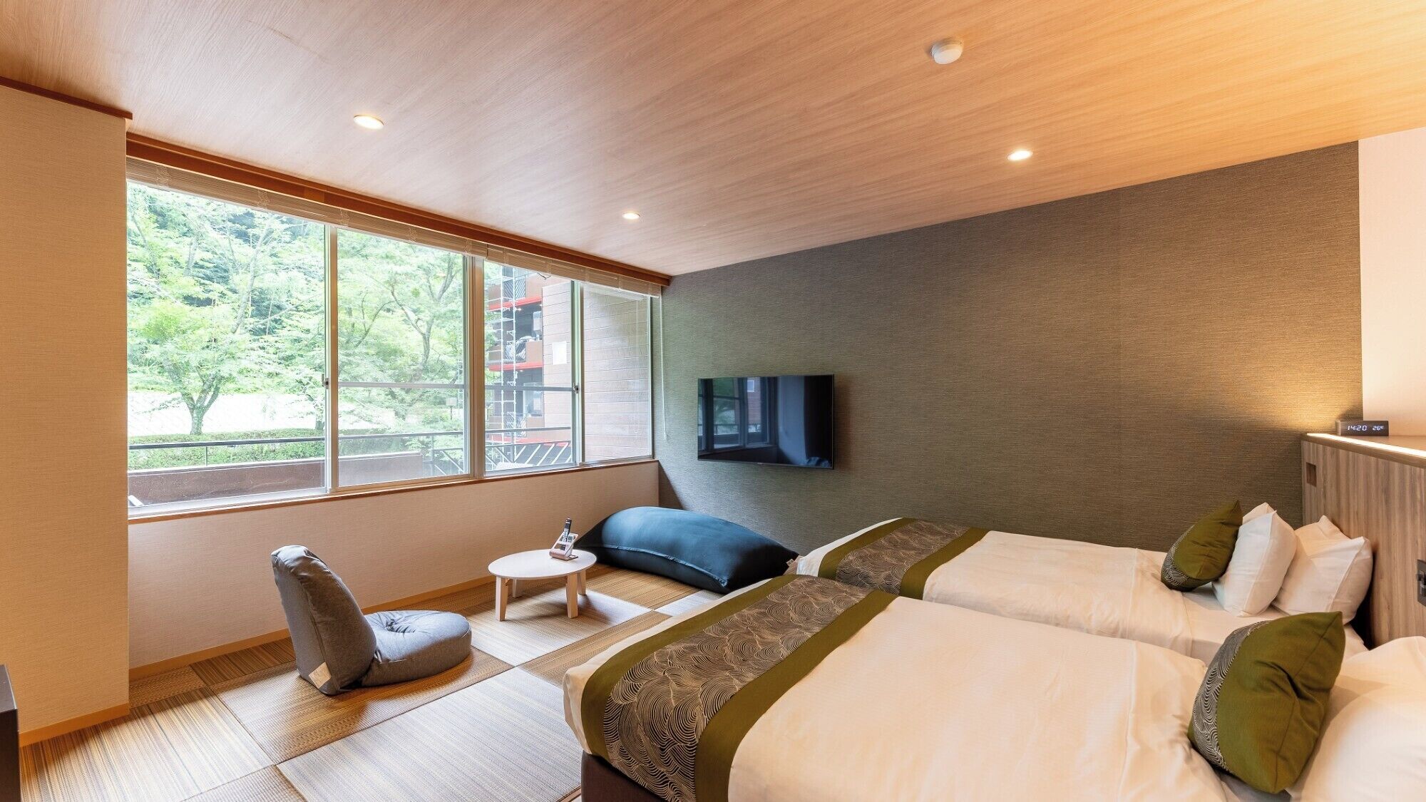 A hotel room with two beds, a TV mounted on the wall, a small table, and a window with a view of trees.